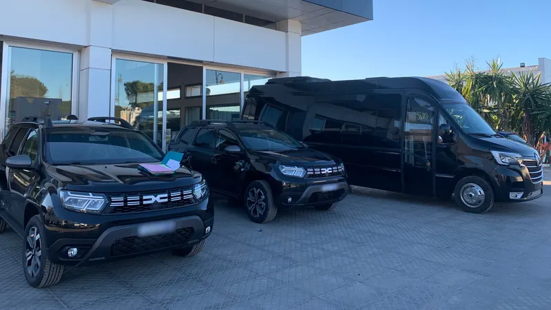 Deux Dacia Duster noirs neufs et un minibus Renault Master noir devant une concession, sous un ciel bleu
