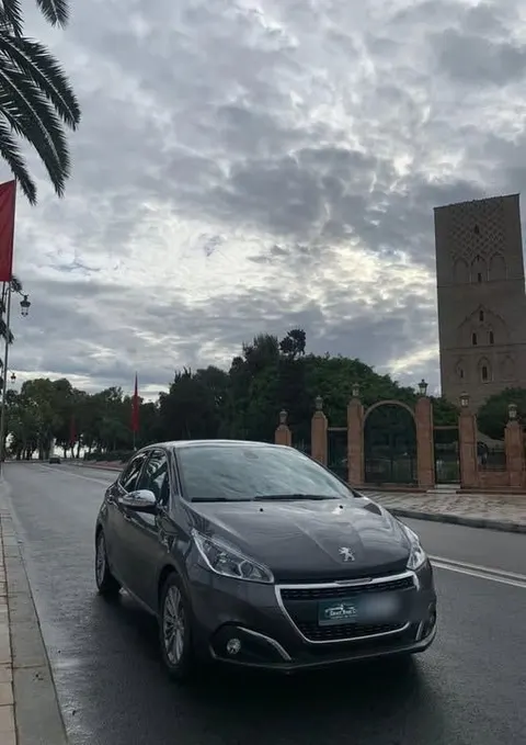 Peugeot 208 grise avec un cadre de plaque Exact Rent Car, garée devant la tour Hassan sous un ciel nuageux