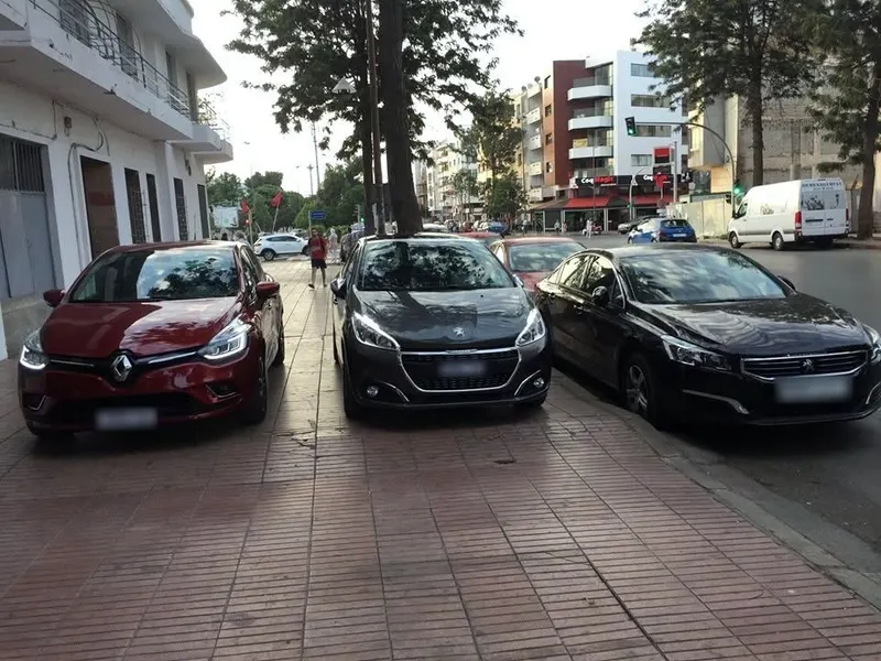 Une Renault Clio rouge et une Peugeot 208 grise de l’agence, garées sur un trottoir arboré de Rabat à côté d’une berline noire